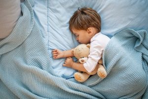 Image of a young boy sleeping on a bed with a teddy bear in his arms.